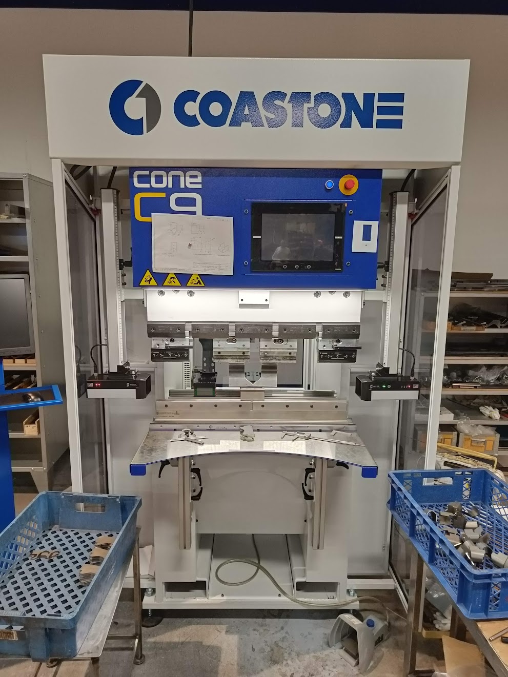 Digitaalinäytöllä ja ohjauspaneelilla varustettu Särmäys eli ohutlevyosien taivutuskone seisoo kahden sinisen, metalliosilla täytetyn säiliön välissä työpajassa.