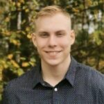 Lyhyet vaaleat hiukset omaava nuori mies, jolla on tummansininen paita, jossa on pieniä valkoisia pisteitä, hymyilee kameralle seisoessaan ulkona sumean vehreyden ja puiden edessä.