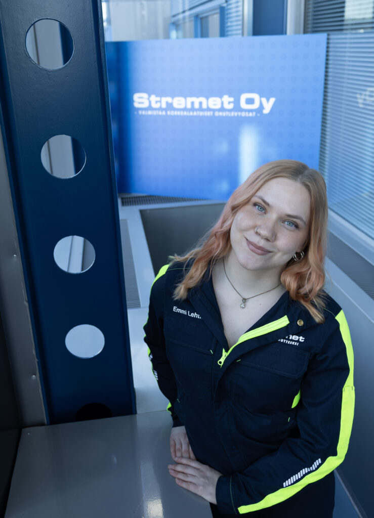 Hymyilevä nainen, jolla on vaaleanpunaiset hiukset, seisoo sisätiloissa yllään tummansininen ja keltainen työasu. Hänen takanaan olevassa sinisessä kyltissä lukee Stremet Oy, ja Yhteystiedot ovat todennäköisesti saatavilla lähellä tässä modernissa teollisuus- tai toimistotilassa.