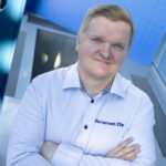Vaaleatukkainen henkilö, jolla on valkoinen paita, jossa on Stremet Oy:n logo ja Yhteystiedot, seisoo sisätiloissa kädet ristissä, katsoo ylöspäin ja hymyilee. Taustalla näkyy sininen kyltti, jossa on Stremet Oy:n logo.