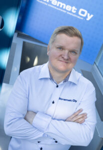 Vaaleatukkainen henkilö, jolla on valkoinen paita, jossa on Stremet Oy:n logo ja Yhteystiedot, seisoo sisätiloissa kädet ristissä, katsoo ylöspäin ja hymyilee. Taustalla näkyy sininen kyltti, jossa on Stremet Oy:n logo.