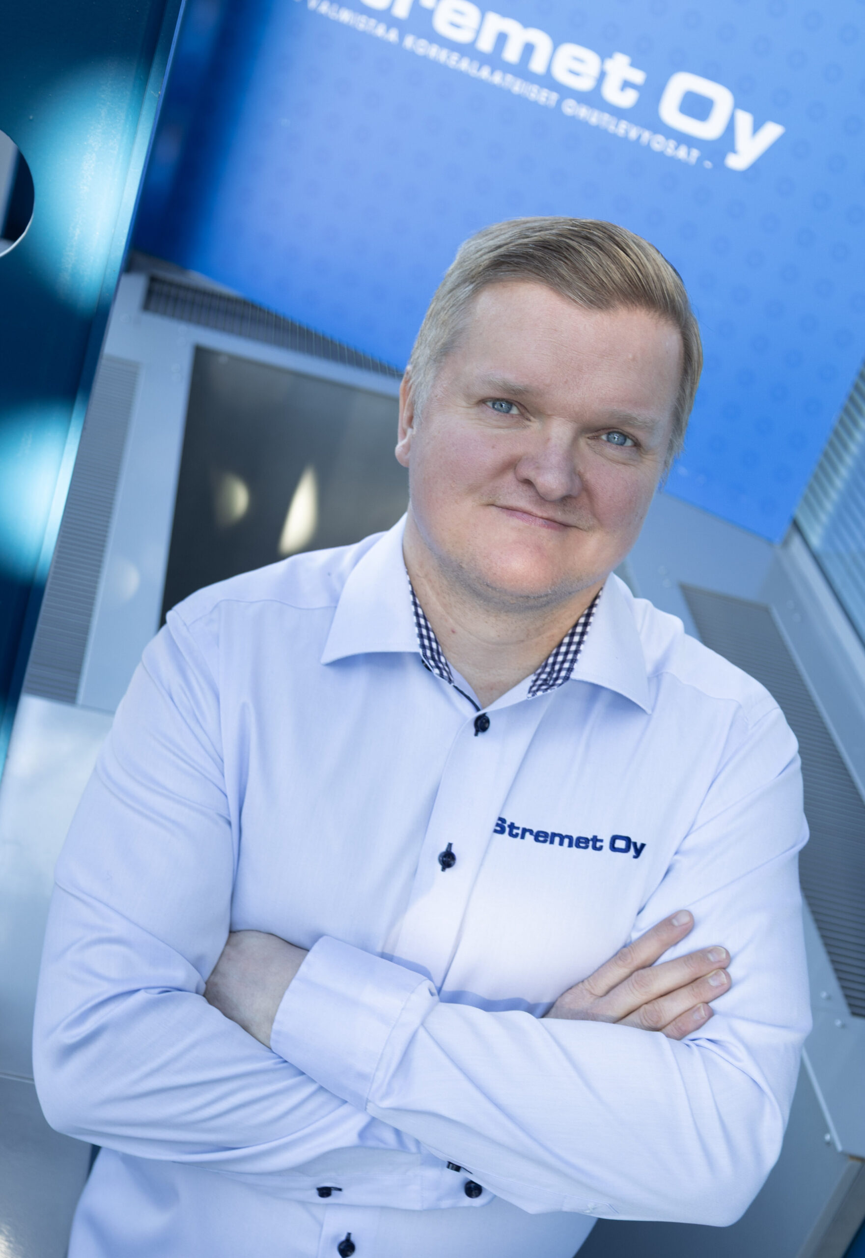 Vaaleatukkainen henkilö, jolla on valkoinen paita, jossa on Stremet Oy:n logo ja Yhteystiedot, seisoo sisätiloissa kädet ristissä, katsoo ylöspäin ja hymyilee. Taustalla näkyy sininen kyltti, jossa on Stremet Oy:n logo.