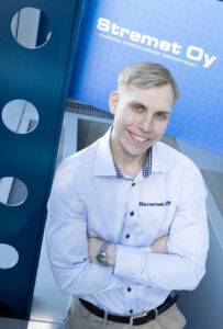 Hymyilevä vaaleahiuksinen mies seisoo kädet ristissä, yllään valkoinen paita, jossa on Stremet Oy:n logo. Taustalla on sininen kyltti, jossa lukee Yhteystiedot ja sama logo sekä suomenkielinen teksti.