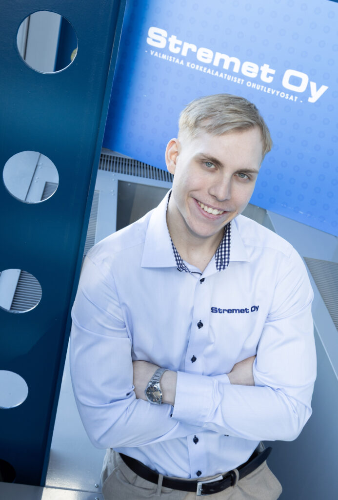Hymyilevä vaaleahiuksinen mies seisoo kädet ristissä, yllään valkoinen paita, jossa on Stremet Oy:n logo. Taustalla on sininen kyltti, jossa lukee Yhteystiedot ja sama logo sekä suomenkielinen teksti.