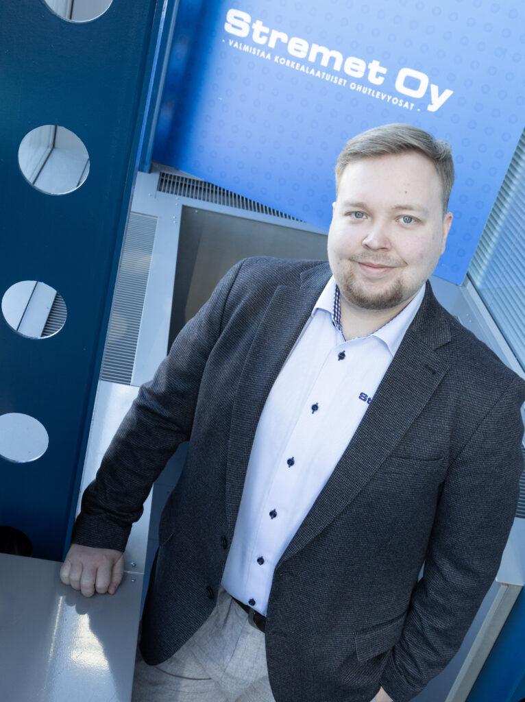 Lyhyet vaaleat hiukset ja leikattu parta omaava mies, jolla on harmaa bleiseri ja valkoinen paita, seisoo sisätiloissa lähellä sinistä seinää, jonka taustalla on Stramet Oy:n kyltti, jossa lukee Yhteystiedot.