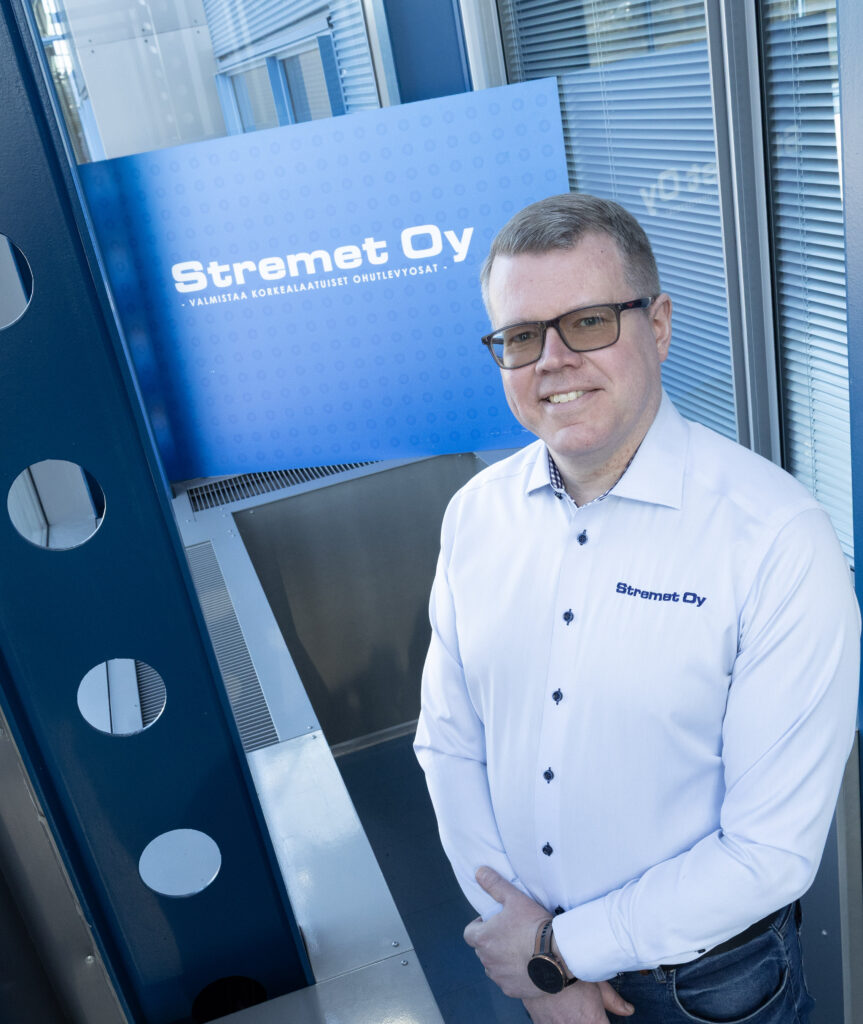 Mies, jolla on silmälasit ja valkoinen paita, jossa on Stremet Oy:n logo, seisoo ja hymyilee lähellä sinistä Yhteystiedot-kylttiä, jossa on sama logo ja suomenkielinen teksti, modernin lasi- ja metallirakennuksen sisällä.