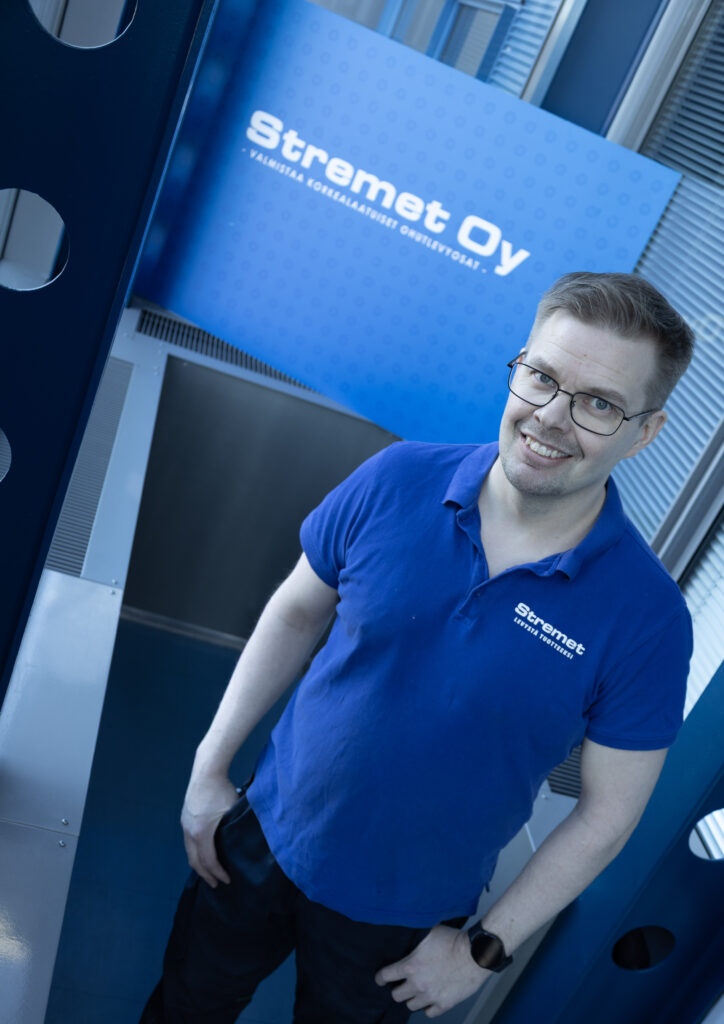 Mies, jolla on silmälasit ja sininen Stremet Oy:n poolopaita, seisoo sisätiloissa ja hymyilee kameralle. Hänen takanaan on sininen kyltti, jossa näkyy Stremet Oy:n logo, suomenkielinen teksti ja Yhteystiedot.