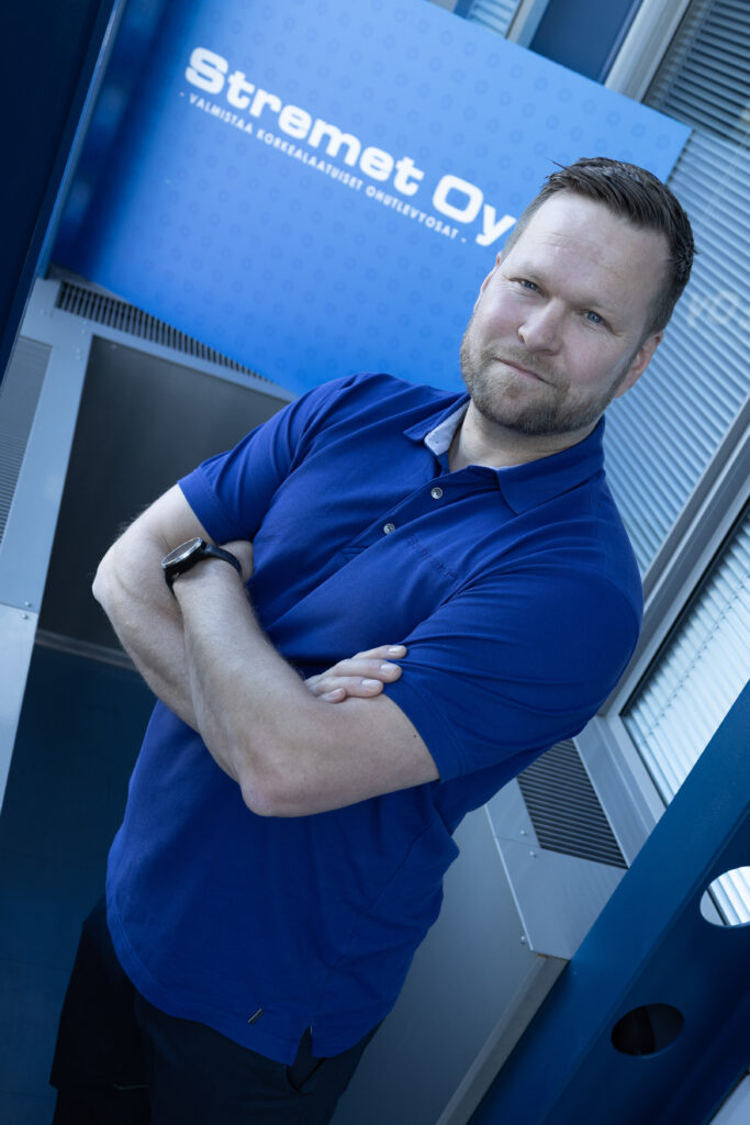 Lyhythiuksinen ja parrakas mies, jolla on sininen poolopaita ja älykello, seisoo kädet ristissä sinisen kyltin edessä, jossa lukee valkoisilla kirjaimilla Stremet Oy ja joka tarjoaa ammattimaiset kasvot Yhteystiedot-sivustolle.