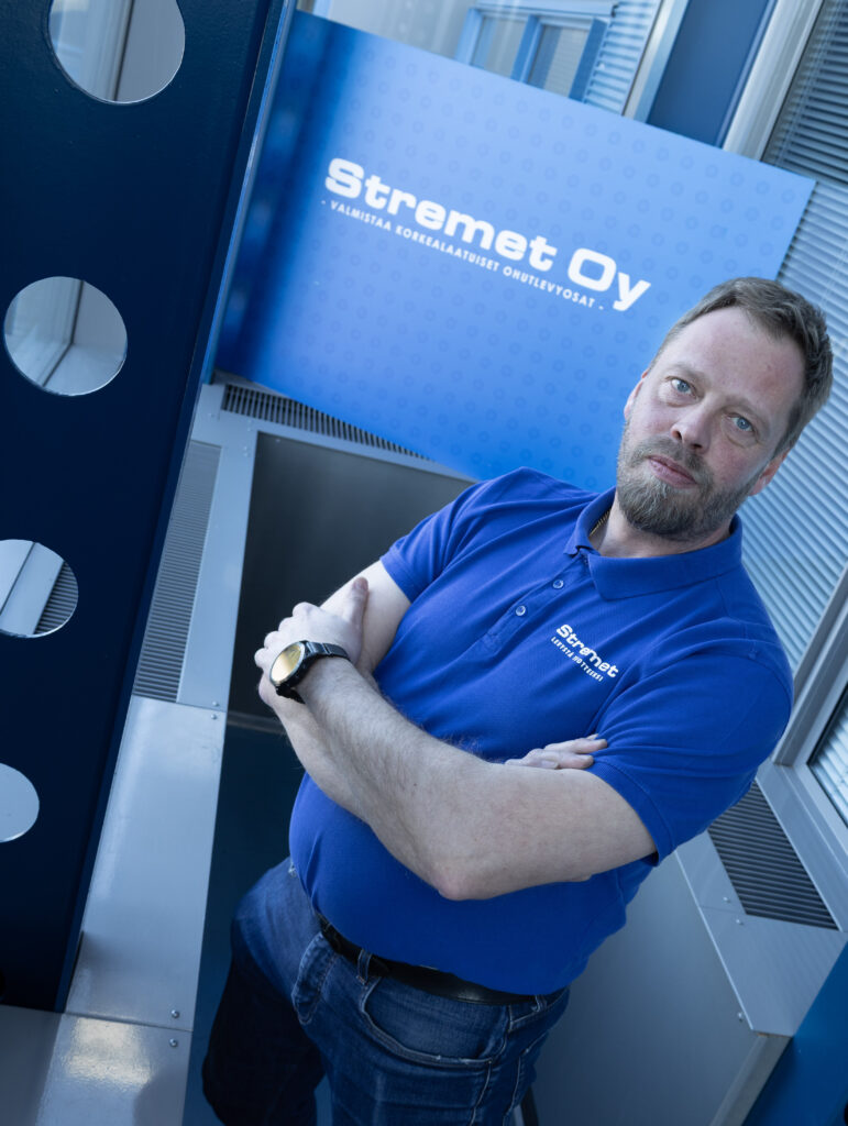Parrakas mies, jolla on sininen Stremet Oy:n poolopaita, seisoo kädet ristissä ja katsoo kameraan. Taustalla näkyy Stremet Oy:n kyltti, joka korostaa yrityksen toimistoa ja Yhteystiedot yksityiskohtia.