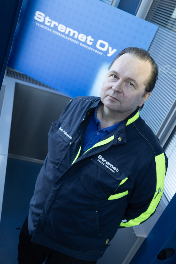 Sinikeltaiseen työtakkiin pukeutunut mies seisoo sinisen Stremet Oy:n kyltin edessä, jossa lukee Yhteystiedot. Hän katsoo kameraan neutraali ilme kasvoillaan modernin teollisuusympäristön sisällä.