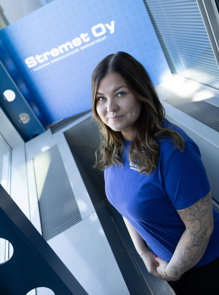 Nainen, jolla on pitkät ruskeat hiukset ja tatuoitu käsivarsi ja joka on pukeutunut siniseen Stremet Oy -t-paitaan, seisoo sisätiloissa suurten ikkunoiden lähellä, ja taustalla näkyy sininen kyltti, jossa on yrityksen nimi, logo ja ”Yhteystiedot”.